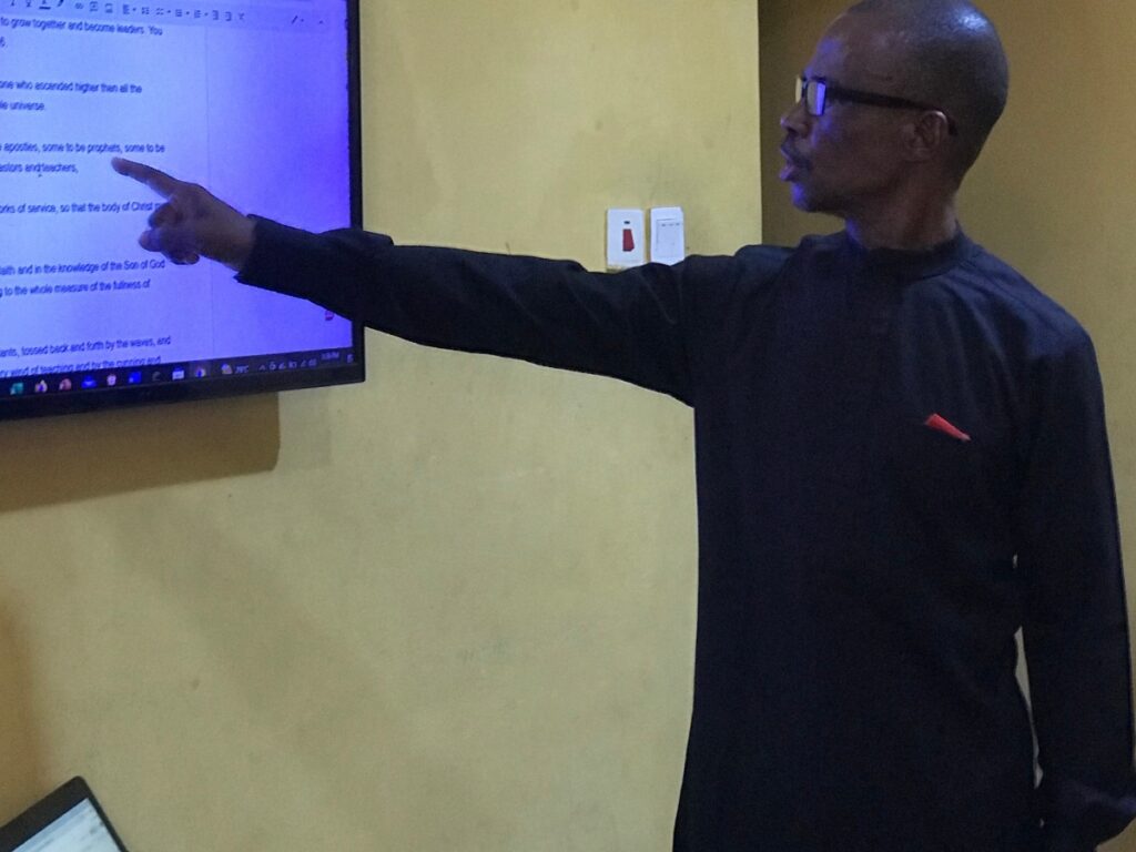 Man teaching in a house church session, pointing to a television screen while explaining a Bible lesson.